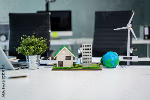 Mini house, office building, globe and wind turbine models on a modern desk, showing sustainable real estate, clean energy and green business ideas.
