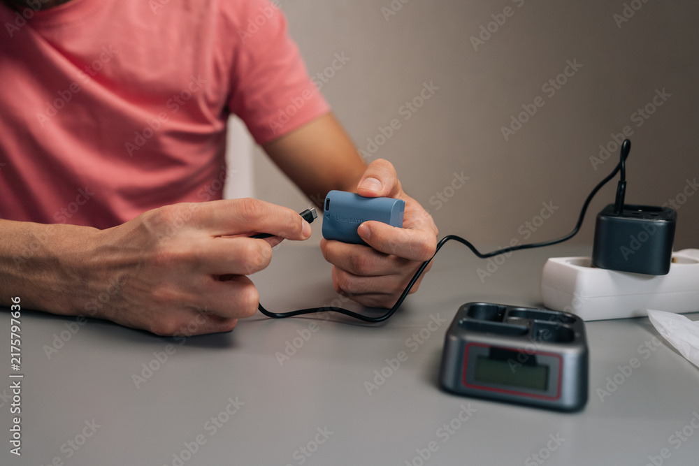 Fototapeta premium Filmmaker plugging lithium-ion battery into multi-slot charging station connected to power strip, ensuring essential camera accessories ready for work, close-up, cropped shot.