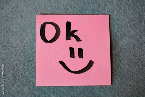 Handwritten word OK with smile icon on square white paper note placed on grey felt background