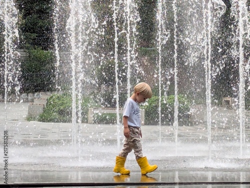 Little boy in yellow wellies standing close to the fountain, laughing as droplets catch the light. Carefree moments of childhood.

