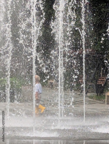A young child in bright wellies, trailing the fountain spray with each step. Giggling as the droplets catch up.

