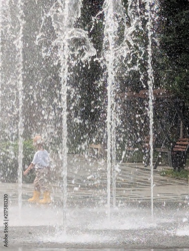 Little boy walking through the light mist of a fountain, yellow boots shining. Following the water's playful path.

