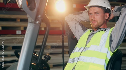 Forklift operator in safety gear relaxes in the drivers seat with hands behind his head during a work break inside an industrial warehouse.