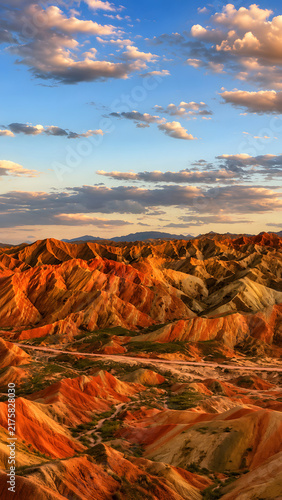 Colorful layered mountains of Zhangye Danxia landform in Gansu, China, glowing in warm sunlight beneath a dramatic blue sky with clouds.