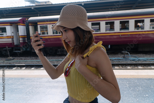 Young Asian female traveler wearing a straw hat and yellow top checking her smartphone while waiting at a railway station platform. Summer vacation and travel lifestyle concept.