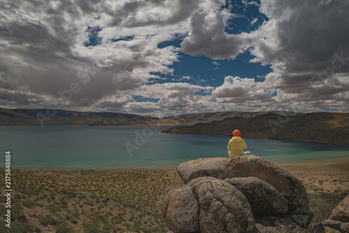 Quadro su tela Rear view of a traveler sitting on a rocky cliff and looking at a scenic turquoi