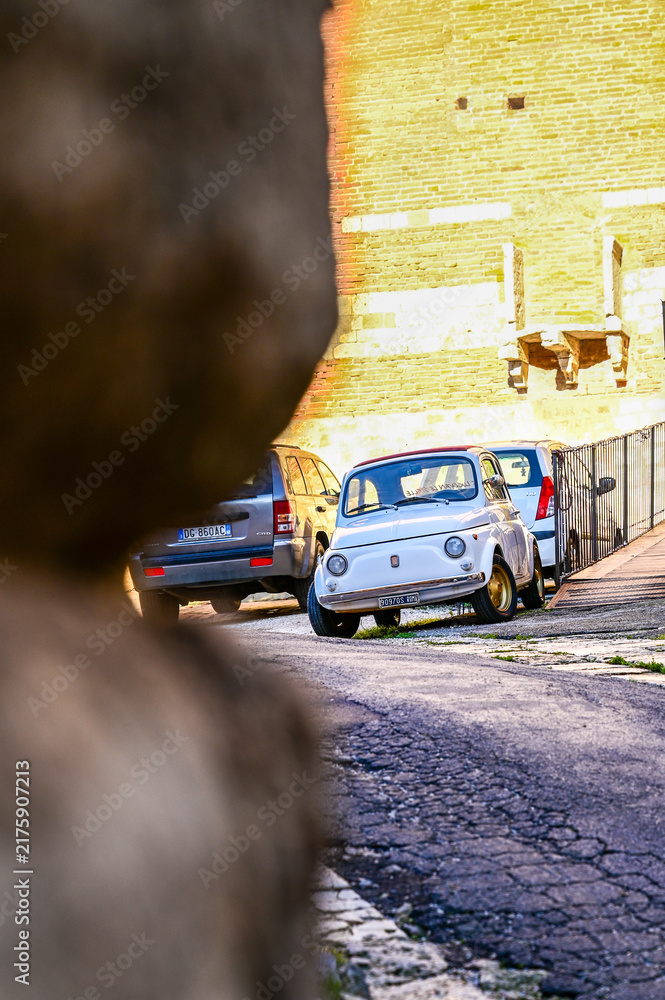 Obraz premium White vintage 1960s Fiat 500 L with a folding roof parked by a historic city wall; a classic Italian cult microcar; Montepulciano, Tuscany, Italy 02.12.2025