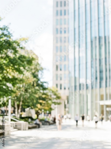 Blurred Modern City Street with Office Buildings, Green Trees, and People Walking on a Sunny Day