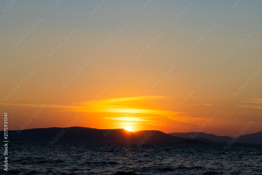 Obraz premium A dramatic sunset over rippling ocean water with the sun partially hidden behind dark mountain silhouettes, casting a vibrant orange and yellow glow across the sky.