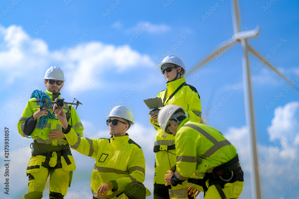 Fototapeta premium Bearded technician holds a grey inspection drone in both hands while a kneeling colleague reaches toward its rotor and a huge white turbine rises in the background