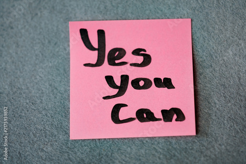 Pink sticky note with handwritten motivational phrase Yes You Can on grey felt background, motivation concept