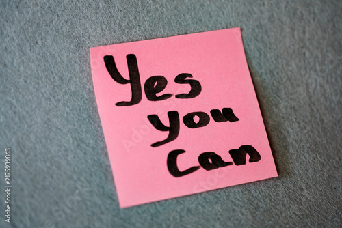 Pink sticky note with handwritten motivational phrase Yes You Can on grey felt background, motivation concept