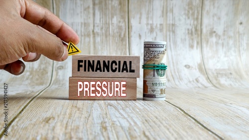 Hand placing a caution sign near blocks reading FINANCIAL PRESSURE beside rolled US dollar bills.