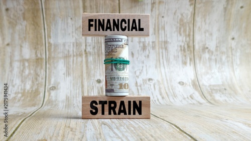 Rolled US dollar bill supporting wooden blocks reading FINANCIAL STRAIN on a light background.