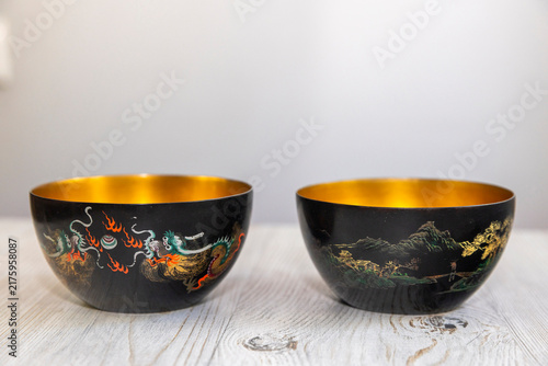 Two black lacquer vintage bowls with gold interiors displayed side by side showing dragon and mountain landscape painted decorations