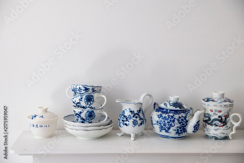 Blue and white ceramic teaware, including a teapot, cups, and a pitcher, arranged on a white shelf against a plain background.
