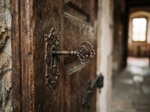 A detailed close-up of an ornate vintage metal key inserted into the keyhole of an antique, weathered wooden door in a stone hallway.