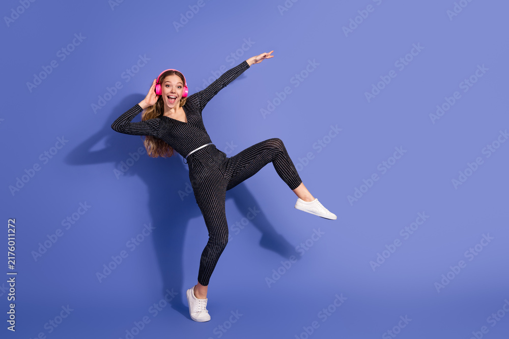Fototapeta premium Young woman in striped jumpsuit wears pink headphones jumping and smiling against a violet background for a stylish fun lifestyle and fashion advertising image
