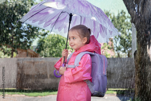 Young schoolgirl in a bright pink raincoat carries a backpack and holds a purple umbrella outdoors on a school day