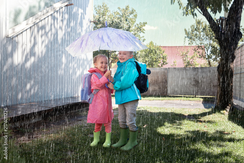 A cheerful boy in a blue raincoat and a girl in a pink raincoat, both with backpacks, share a purple floral umbrella, laughing in the autumn rain. Candid.
