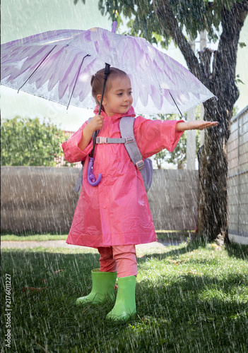 Happy toddler wearing pink raincoat, rubber boots and backpack standing under umbrella and playing with water drops in rainy day in a grassy garden