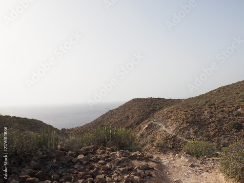 Fotografía Narrow dirt path leading through a rocky mountain pass towards the sea