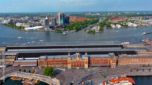 Aerial drone view of Amsterdam Centraal railway station with trams and travelers on Stationsplein, platform roofs, ferries on the IJ river and the Amsterdam Noord skyline, Netherlands.
