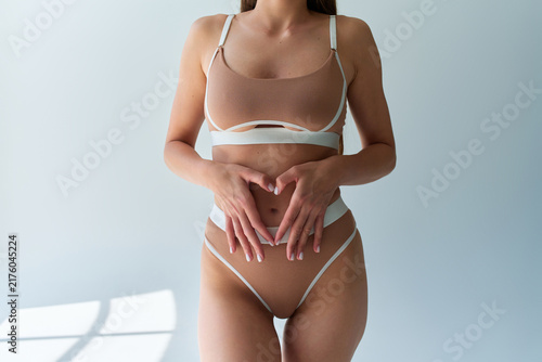 Cropped woman in beige lingerie touching her abdomen in a bright minimalist room, representing body care, confidence, wellness, and healthy lifestyle.
