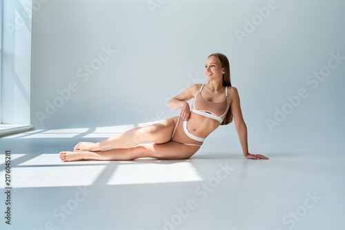 Smiling woman in beige lingerie sitting on a bright studio floor, relaxing in sunlight and window shadows against a clean minimal background.