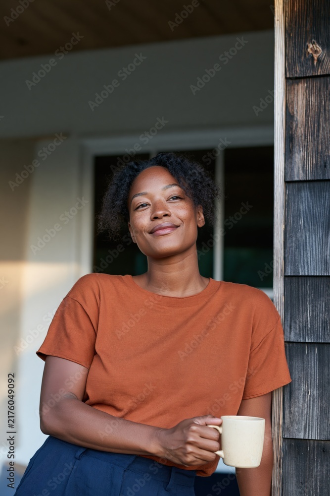 Obraz premium Relaxed woman leaning on a porch post with a mug wearing an orange tee casual lifestyle portrait for social media and advertising