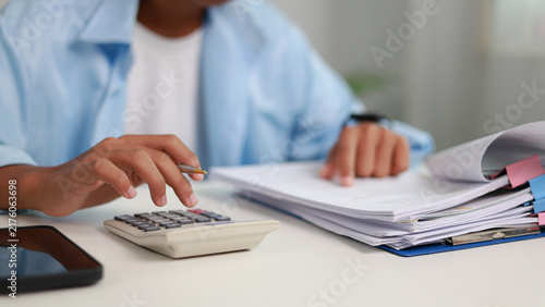 Close up male accountant hand pressing calculator button and checking financial document paperwork with stack of papers on desk. Tax invoice review and budget audit analysis concept.