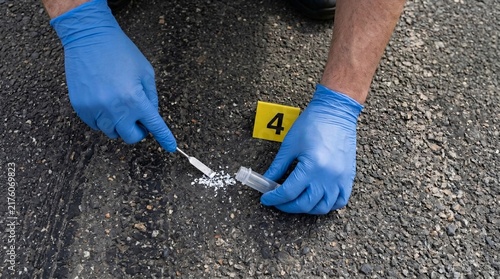Overhead close-up of a forensic scientist wearing blue gloves using a spatula to collect a white powder sample into a container near evidence marker 4.