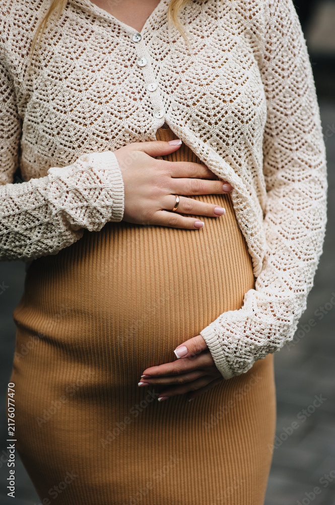 Naklejka premium A close-up photo of a young pregnant woman in a dress, gently holding her large belly in her hands. Waiting for a miracle. Pregnancy concept.