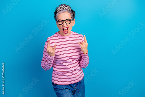 Senior woman wearing a tiara and glasses making rock hand signs and shouting with playful rebellious energy in a pink striped shirt on a bright blue background