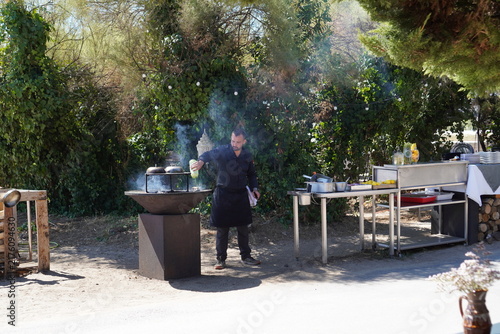 Картината върху платно A chef prepares food at an outdoor stainless steel station beside a smoking braziers