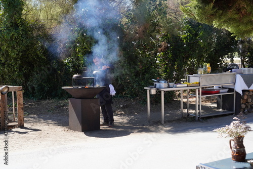 Картината върху платно A chef prepares food at an outdoor stainless steel station beside a smoking braziers