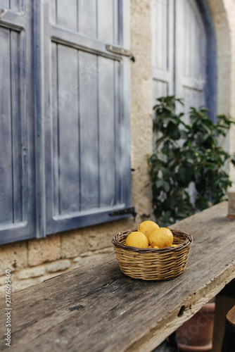 Fresh Lemons in Wicker Basket on Rustic Table. Rustic still life with fresh lemons in a wicker basket, weathered wooden table and blue shutters of a traditional stone house, Alacati, Turkiye, Aegean. 