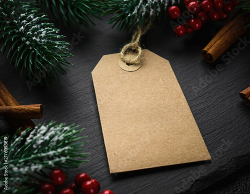 Blank kraft paper gift tag surrounded by festive Christmas decorations including snowy pine branches, bright red berries, and aromatic cinnamon sticks on a dark background