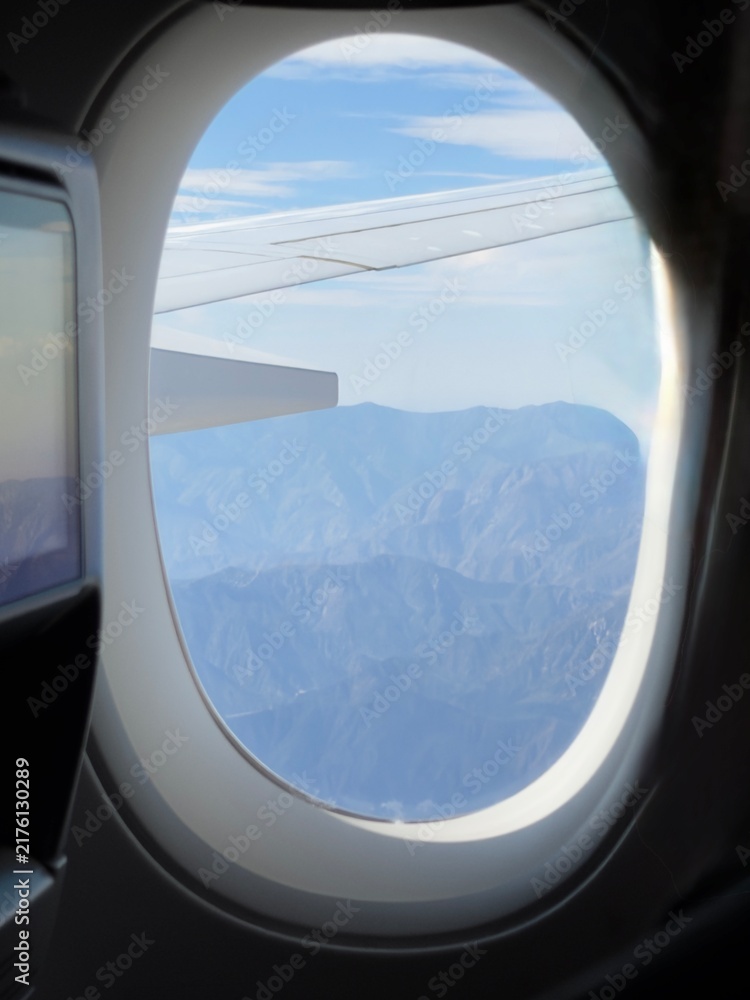 Fototapeta premium Airplane wing flying above rugged mountains with blue sky seen from passenger window