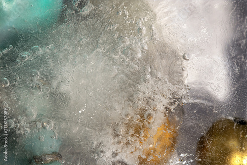Close-up of transparent ice with trapped air bubbles and blurred turquoise and golden objects frozen inside.