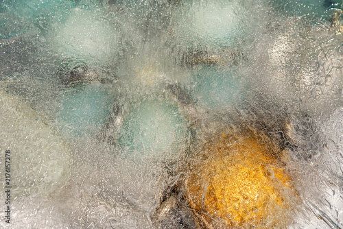 Close-up of transparent ice with trapped air bubbles and blurred turquoise and golden objects frozen inside.