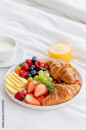 Cozy breakfast in bed with croissants, fresh berries, grapes, cheese, coffee and orange juice on white bedding