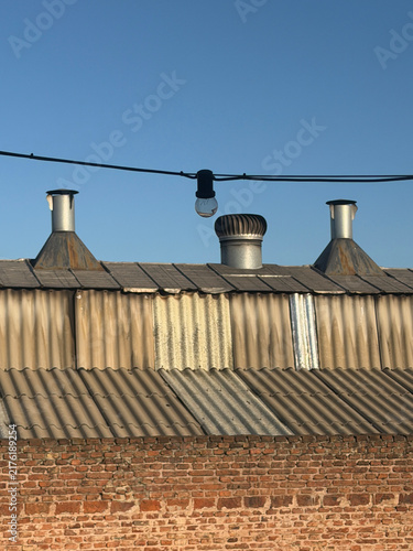 Industrial rooftop with metal chimneys, weathered corrugated roofing and brick wall under a clear blue sky.
