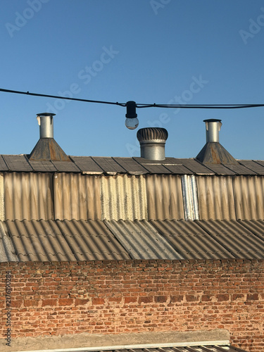 Industrial rooftop with metal chimneys, weathered corrugated roofing and brick wall under a clear blue sky.