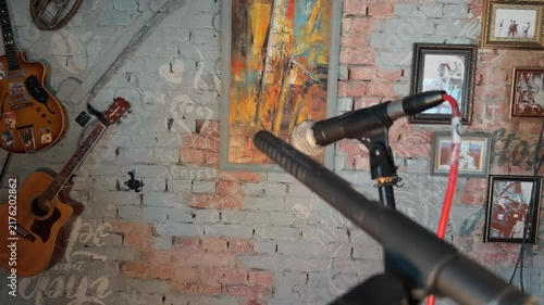 Professional broadcasting microphones on stands with red audio cable in foreground inside vintage recording studio with painted brick wall decorated by old art and acoustic guitars handheld arc shot