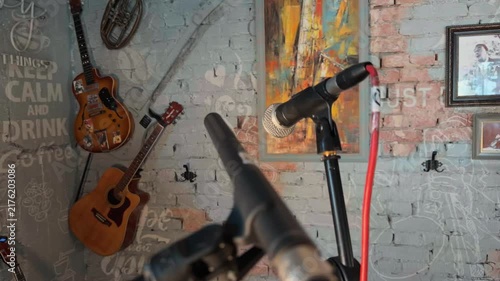 Professional microphones on stands ready for live music performance on empty stage inside cozy retro comedy club with painted brick wall vintage guitars and neon sign handheld orbiting arc shot