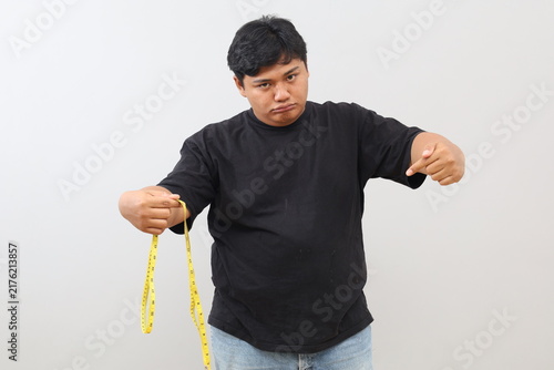 Konstfotografi An overweight man holds a measuring tape while pointing at his belly with a sulky expression, standing isolated against a white background