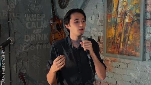 Emotional Young Asian Man Storytelling Into Microphone During Soulful Performance on Stage in Cozy Cafe Club with Guitars and Brick Wall. Medium Handheld Candid Shot and Warm Atmosphere.