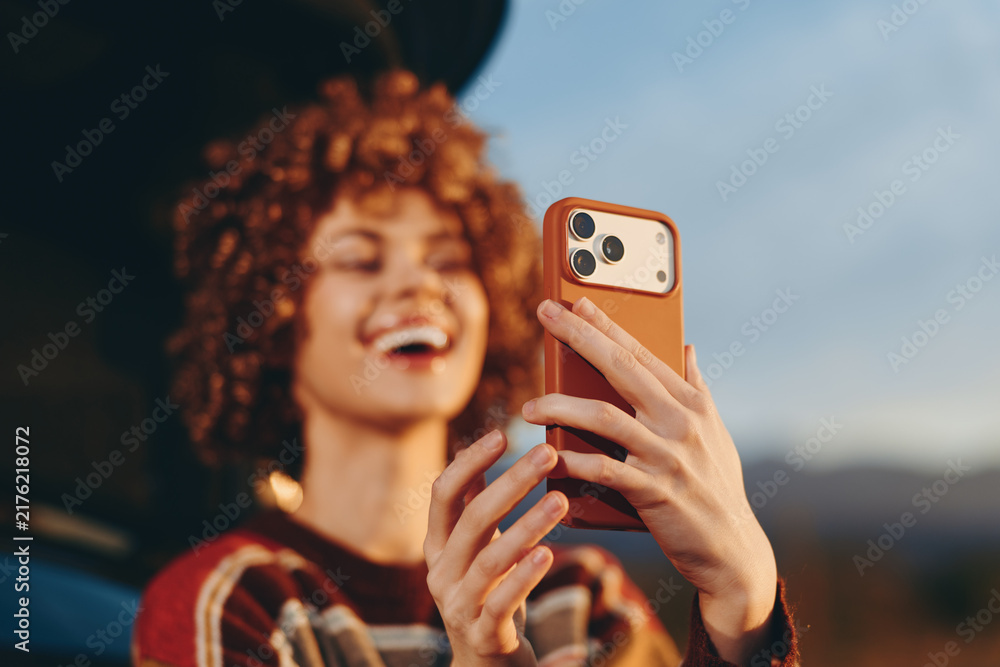 Fototapeta premium Woman selfie smartphone lifestyle outdoors portrait sunset closeup, young adult smiling, curly hair and freckles, holding phone for camera, warm golden hour light, blurred mountains background