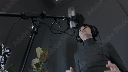 Focused young Asian male voiceover actor putting on black headphones preparing for commercial recording session in modern dark home studio looking at condenser microphone low angle authentic shot
